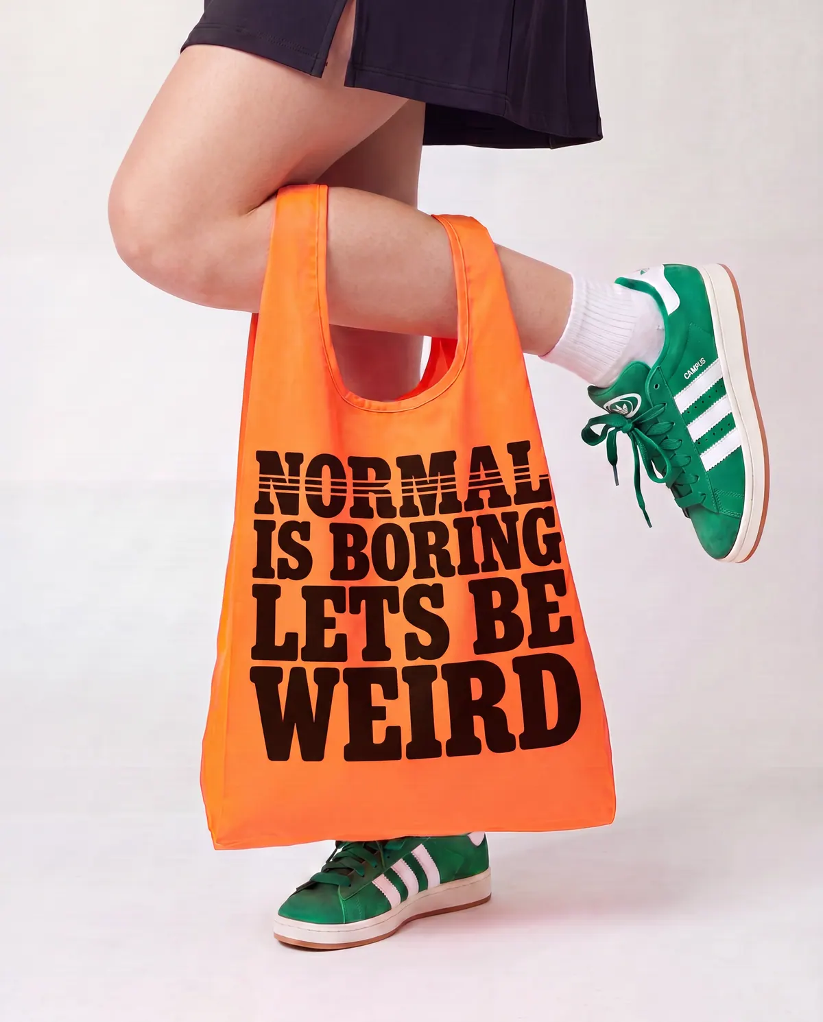 Un tote bag orange marqué « Normal is boring, let's be weird », tenu au bout du bras au-dessus de baskets vertes.