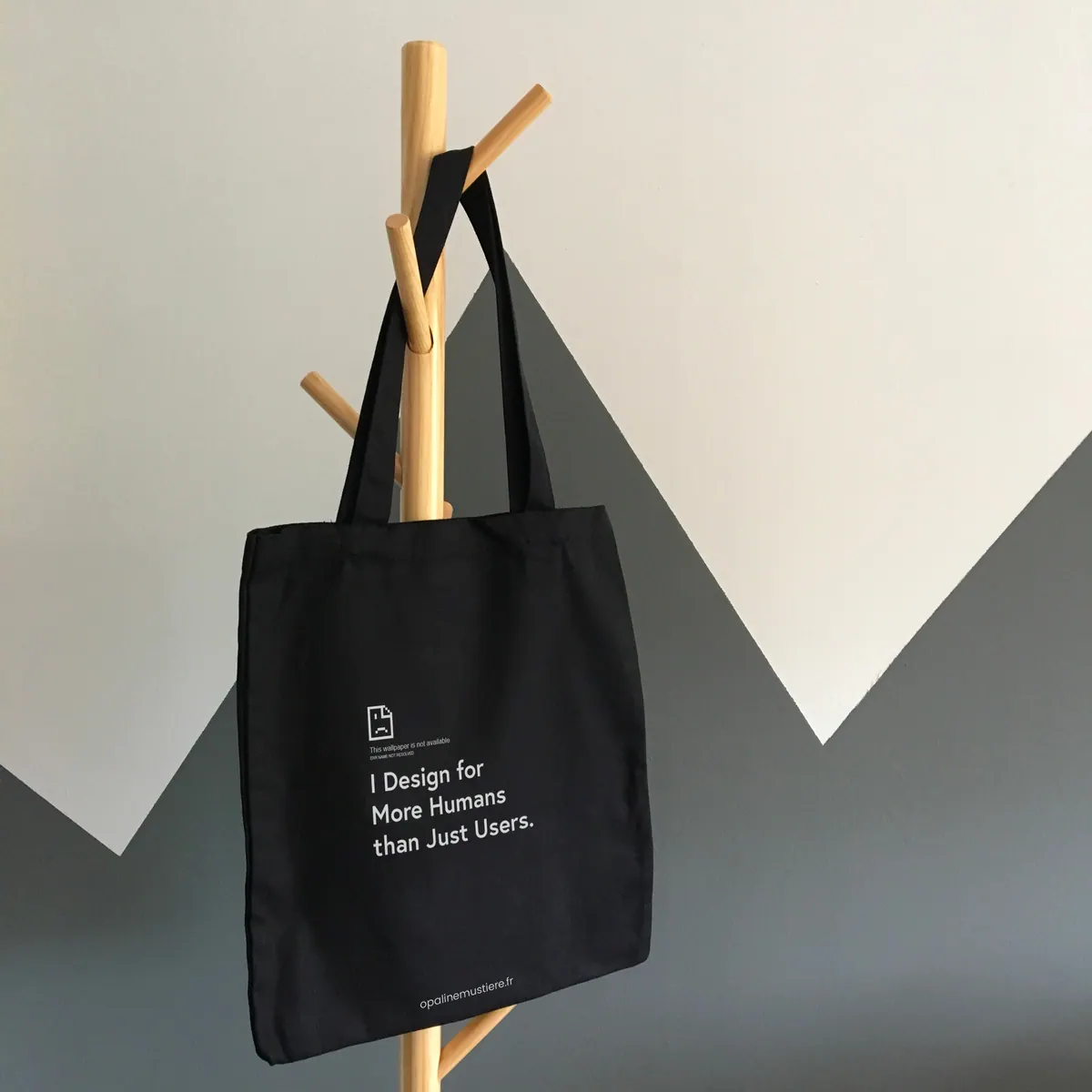 Un tote bag noir sur un portemanteau, imprimé « I design for more humans than just users ».